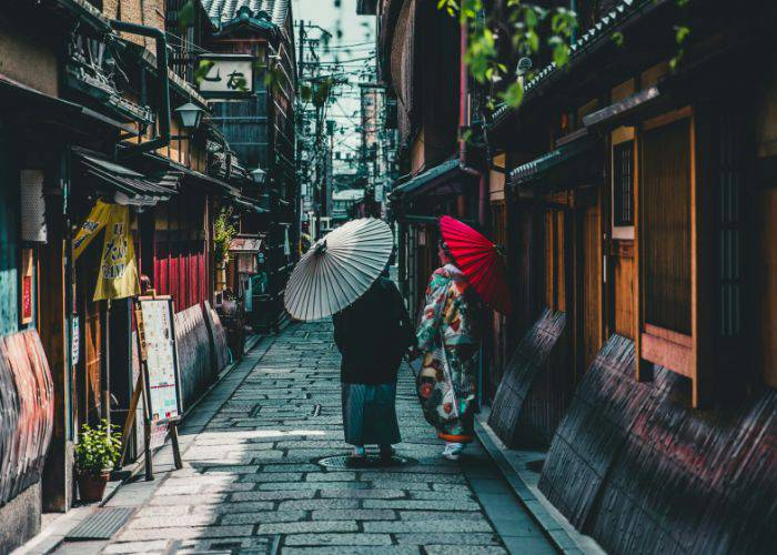 Two people wearing kimono wandering down the traditional streets of Kyoto, holding umbrellas.